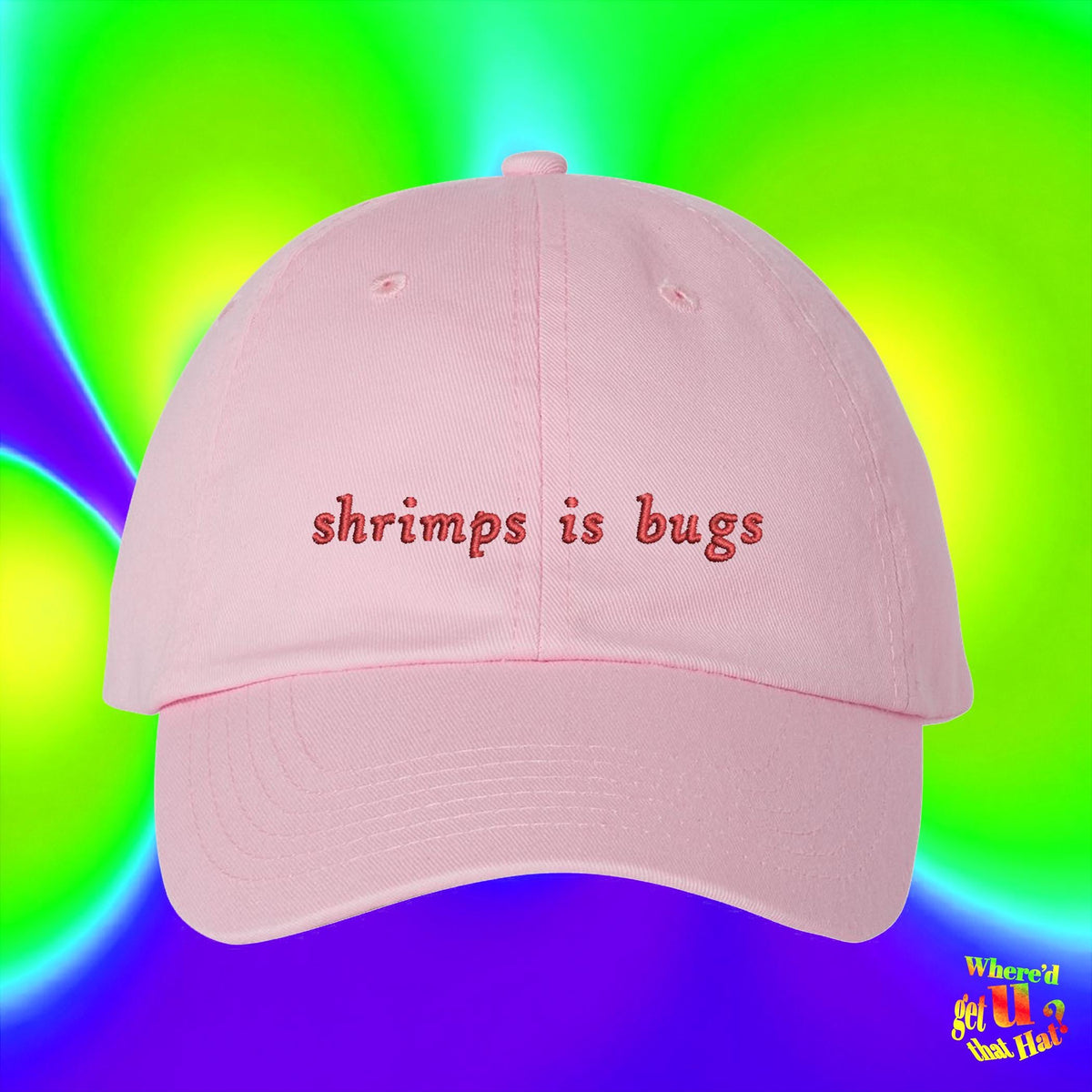 A pink baseball cap with the text "shrimps is bugs" printed on it, against a colorful background.